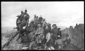 Men and women in early 20th century hiking clothes on a mountain peak with other peaks in the background